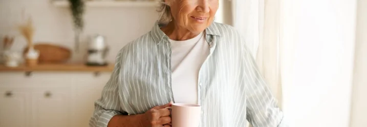 Ältere Person mit einer Tasse blickt lächelnd aus dem Fenster.